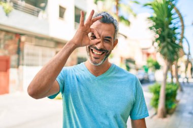 Orta yaşlı, kır saçlı, günlük kıyafetler giyen, gülümseyen, mutlu, el pençe divan duran bir adam. 