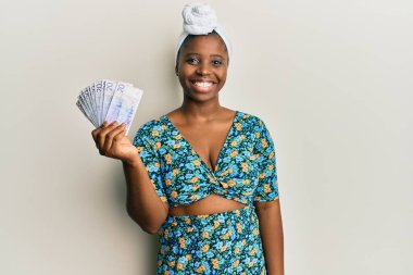 Saç sarığı takan genç Afrikalı bir kadın elinde 20 İsveç kronu banknotuyla pozitif ve mutlu görünüyor ve gülümseyerek dişlerini gösteriyor. 