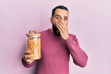 Sakallı genç adam elinde makarna kavanozuyla ağzını eliyle kapadı, şok oldu ve hata yapmaktan korktu. şaşırmış ifade 