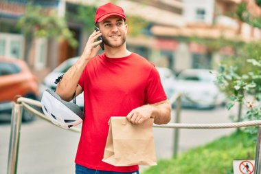 Beyaz atlı genç teslimatçı akıllı telefondan konuşuyor ve şehirde kağıt torba taşıyor..