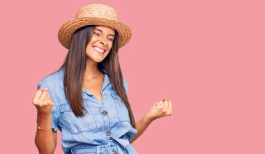 Genç İspanyol kadın yaz şapkası takıyor. Çok mutlu ve heyecanlı. Kolları havada, gülümseyen ve başarı için bağıran kazanan jestler yapıyor. kutlama konsepti. 