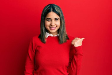 Gündelik kıyafetler giyen, gülümseyen, gülümseyen ve başparmağıyla yan tarafı gösteren genç Latin kadın.. 