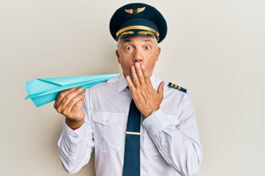 Handsome middle age mature man wearing airplane pilot uniform holding paper plane covering mouth with hand, shocked and afraid for mistake. surprised expression 