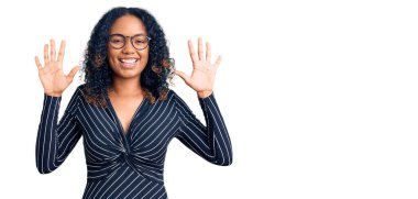 Genç Afro-Amerikalı kadın günlük kıyafetler ve gözlük takıyor ve parmaklarıyla on numarayı gösterirken kendinden emin ve mutlu gülümsüyor.. 