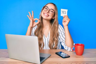 Genç, sarışın bir kadın elinde evet hatırlatıcısıyla masada oturmuş parmaklarıyla işaret veriyor, gülümsüyor ve mükemmel bir sembol oluşturuyor. 