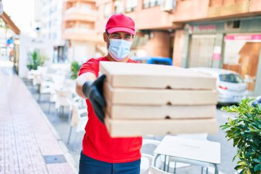 Üniforma giyen ve koronavirüs koruma maskesi takan genç teslimatçı. Kasaba caddesinde pizza kutularını teslim ediyorum..