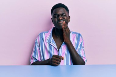 Genç Afro-Amerikan erkek, diş ağrısı ya da diş hastalığından dolayı masaya oturmuş ağzıyla dokunuyor ve acı dolu bir ifade takınıyor. dişçi 