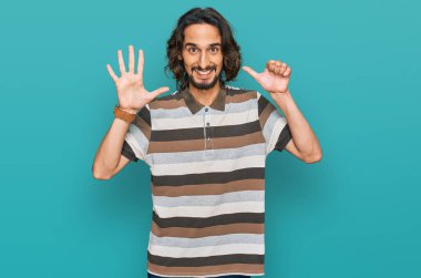 Latin asıllı genç adam gündelik giysiler giyip altı numaralı parmaklarıyla işaret ederken kendinden emin ve mutlu bir şekilde gülümsüyor.. 