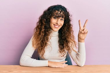 Latin kökenli genç bir kız, gündelik giysiler içinde masada oturuyor ve iki numaralı parmaklarıyla işaret ediyor. Kendinden emin ve mutlu gülümsüyor.. 