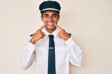 Uçak pilotu üniforması giyen genç İspanyol adam ağzı açık gülümsüyor, parmakları işaret ediyor ve neşeli bir gülümseme sergiliyor. 