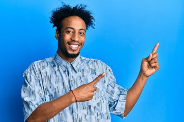 Sakallı, sıradan giyinmiş, gülümseyen ve kameraya bakan iki eliyle ve parmaklarıyla yüzünü gösteren Afrikalı genç bir Amerikalı.. 