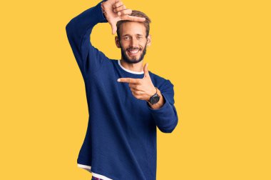 Yakışıklı, sakallı, sıradan bir süveter giyen, gülümseyen, elleri ve parmakları mutlu yüzlü sarışın bir adam. yaratıcılık ve fotoğrafçılık kavramı. 