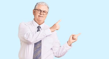 İş kıyafetleri giymiş, gülümseyen ve kameraya bakan yaşlı, gri saçlı bir adam. İki eli ve parmakları yanda.. 