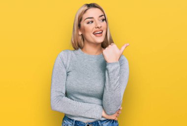 Gündelik kıyafetler giyen, mutlu yüzlü gülümseyen ve baş parmağıyla yan tarafı gösteren beyaz bir kadın.. 