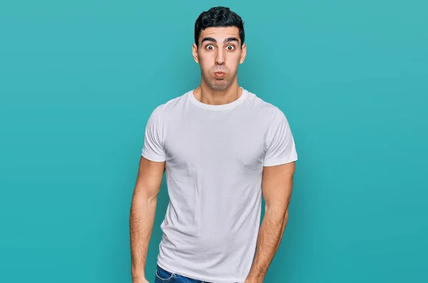 カジュアルな白いTシャツを着たハンサムなヒスパニック系の男が面白い顔で頬をパフ 口は空気で膨らみ狂気の表情で — ストック写真