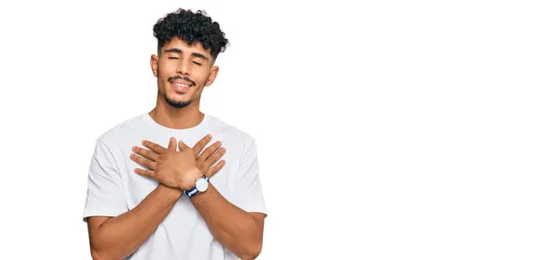 年轻的阿拉伯男子穿着随意的白色T恤 双手放在胸前笑着 闭着眼睛 脸上挂着感激的手势 健康概念 — 图库照片