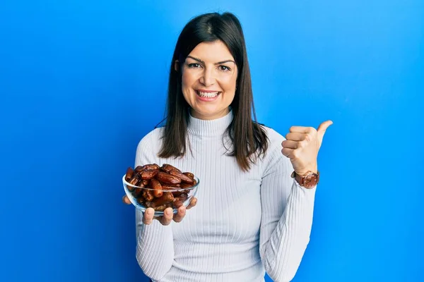 Mujer Hispana Joven Sosteniendo Dátiles Bowl Apuntando Pulgar Hacia Lado —  Fotos de Stock