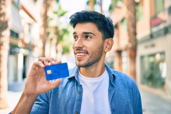 Young latin man smiling happy holding credit card walking at the city.