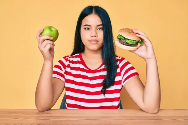 Młoda Azjatka Porównująca Hamburgera Zdrowe Jabłko Zrelaksowana Poważnym Wyrazem Twarzy — Zdjęcie stockowe