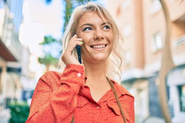 Genç sarışın kadın gülümsüyor. Şehirdeki akıllı telefondan konuşuyor..