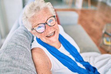 Yaşlı, yaşlı, gri saçlı, mutlu bir kadın. Evdeki kanepede oturuyor.