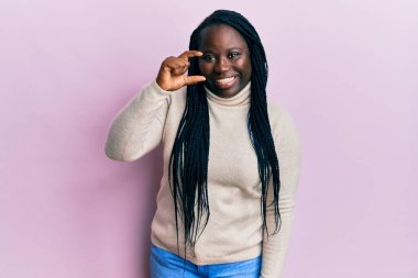 Saçları örgülü, sıradan bir kazak giyen, gülümseyen, kendine güvenen, el hareketi yapan, parmaklarıyla işaret yapan ve kameraya bakan genç siyahi bir kadın. ölçüm kavramı. 