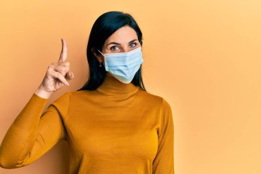 Tıbbi maske takan genç beyaz bir kadın bir fikir veya soruyla gülümsüyor ve mutlu bir yüzle işaret ediyor. 