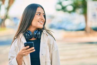 Genç İspanyol kız şehirde akıllı telefon ve kulaklık kullanarak mutlu bir şekilde gülümsüyor..