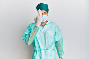 Young handsome man wearing surgeon uniform and medical mask covering one eye with hand, confident smile on face and surprise emotion. 