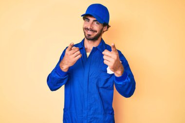 Kıvırcık saçlı, ayı kılıklı, işçi tulumu giyen yakışıklı genç adam mutlu ve komik yüzüyle kamerayı işaret ediyor. iyi enerji ve titreşimler. 