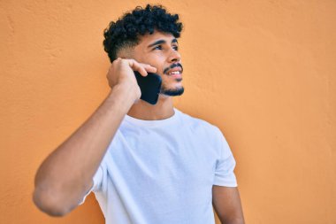 Genç Arap adam gülümsüyor. Akıllı telefonuyla konuşuyor. Duvara yaslanıyor..