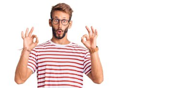 Sakallı, sıradan kıyafetler ve gözlük takan yakışıklı sarışın. Şaşırmış ve şok olmuş bir şekilde parmaklarıyla onay sembolü yapıyor. çılgın ifade 