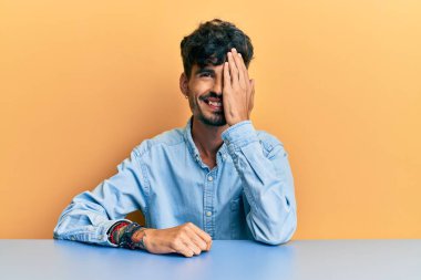 Latin asıllı genç adam gündelik giysiler içinde masada oturuyor bir gözünü eliyle kapatıyor, yüzünde kendine güvenen bir gülümseme ve sürpriz bir duygu var.. 