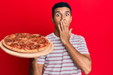 Genç Arap adam İtalyan pizzasını elinde tutarken ağzını eliyle kapadı, şok oldu ve hata yapmaktan korktu. şaşırmış ifade 