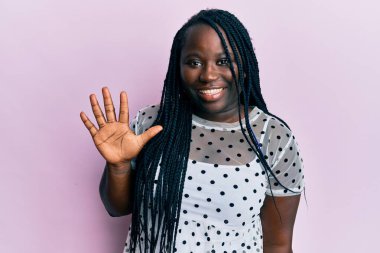 Saçları örgülü genç siyahi bir kadın beş numarayı gösterip gösterirken kendinden emin ve mutlu bir şekilde gülümsüyor.. 