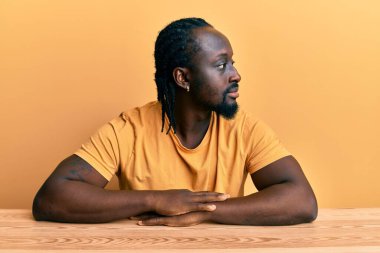 Handsome young black man wearing casual clothes sitting on the table looking to side, relax profile pose with natural face with confident smile. 