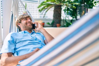 Orta yaşlı yakışıklı bir adam evinin terasında, bir hamakta uzanıp telefonda konuşuyor.