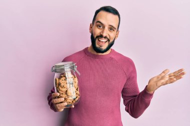 Sakallı genç adam elinde çikolatalı kurabiye kavanozuyla başarıyı mutlu bir gülümsemeyle kutluyor ve kazanan el kaldırarak ifade ediyor. 