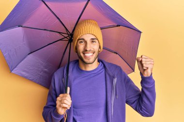 Young handsome man holding umbrella screaming proud, celebrating victory and success very excited with raised arm 
