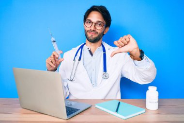 Handsome hispanic doctor man at the clinic holding syringe with angry face, negative sign showing dislike with thumbs down, rejection concept 
