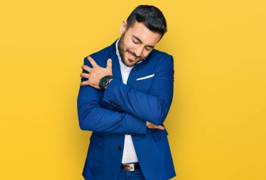 Genç İspanyol bir adam iş ceketi giyiyor mutlu ve olumlu bir şekilde kendine sarılıyor, kendinden emin gülümsüyor. Sevgi ve özgüven. 