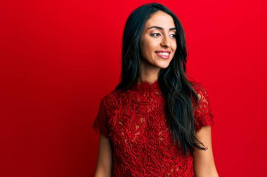 Güzel İspanyol bir kadın, kırmızı arka planda zarif kıyafetler giyip yüzünde gülümseme, doğal bir ifade ile yan yana bakıyor. Kendinden emin gülen. 
