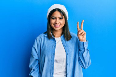 Şirin yün şapkalı genç Latin kadın iki numaralı parmaklarıyla kendini beğenmiş ve mutlu bir şekilde gülümsedi.. 