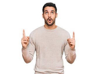 Gündelik giysiler giyen genç İspanyol adam şaşırmış ve şaşırmış bir şekilde yukarı bakıyor ve parmaklarıyla işaret ediyor ve kollarını kaldırıyor.. 