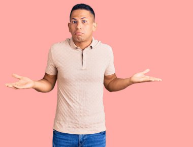 Gündelik kıyafetler giyen genç, yakışıklı Latin bir adam. Kolları ve elleri havada, kafası karışmış bir ifade. şüphe kavramı. 