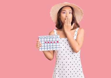 Yaz şapkası takan genç, güzel bir Çinli kız. Elinde kalp takvimi var. Ağzı eliyle kapatıyor. şaşırmış ifade 