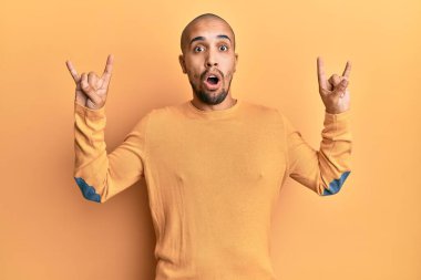 İspanyol yetişkin bir adam sarı arka planda rock hareketi yapıyor. Şaşırmış ve şaşırmış bir ifade, korku ve heyecanlı bir yüz ifadesi var.. 