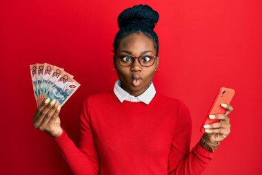 Young african american woman using smartphone holding australian dollar banknotes making fish face with mouth and squinting eyes, crazy and comical. 