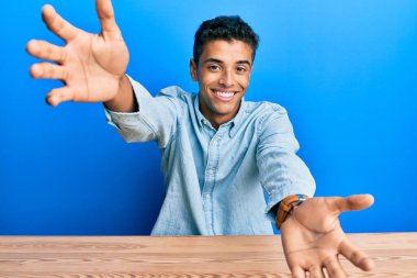 Genç, yakışıklı, Afro-Amerikalı, gündelik giysiler içinde masaya oturmuş kameraya bakıyor ve sarılmak için kollarını açmış gülümsüyor. Mutluluğu kucaklayan neşeli bir ifade.. 