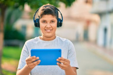 Sevimli Latin çocuk mutlu bir şekilde gülüyor. Parkta kulaklık ve dokunmatik ped kullanıyor..